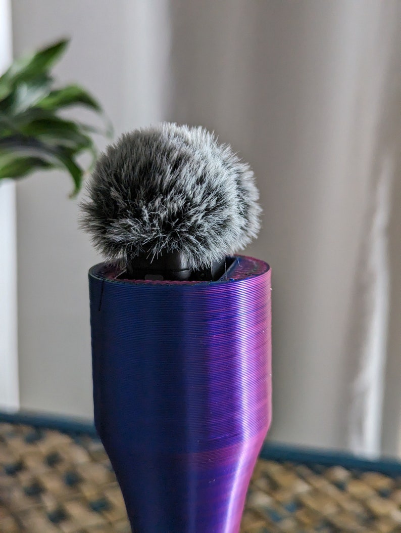 May include: A microphone with a fluffy, gray windscreen sits atop a cylindrical, gradient-colored stand. The stand transitions from blue to purple, with visible layered texture. The background includes a blurred plant and a neutral-toned wall.
