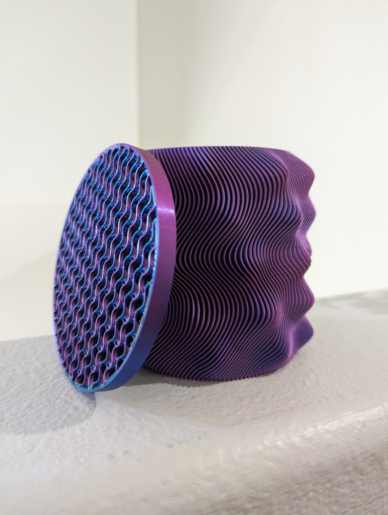 Op de afbeelding: Een 3D-geprinte, abstracte sculptuur in paarse en blauwe tinten. Het hoofdonderdeel heeft een golvende, geribbelde textuur, terwijl het losstaande cirkelvormige deel een patroon met een roosterachtig ontwerp heeft. Het stuk staat tegen een witte achtergrond.
