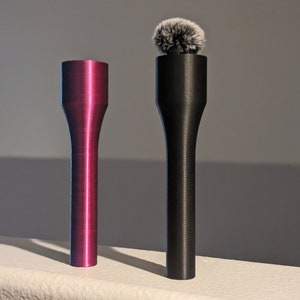 May include: Two microphone handles, one magenta and one black. The magenta handle is smooth and cylindrical with a flared top. The black handle has a fluffy microphone cover on top. Both are standing on a white surface.