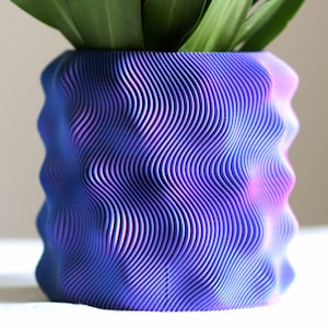 Puede incluir: Una maceta azul y morada con un patr&oacute;n ondulado y una planta en su interior. La maceta tiene un dise&ntilde;o texturizado &uacute;nico con l&iacute;neas verticales que crean una apariencia de onda. La planta tiene hojas largas y verdes.