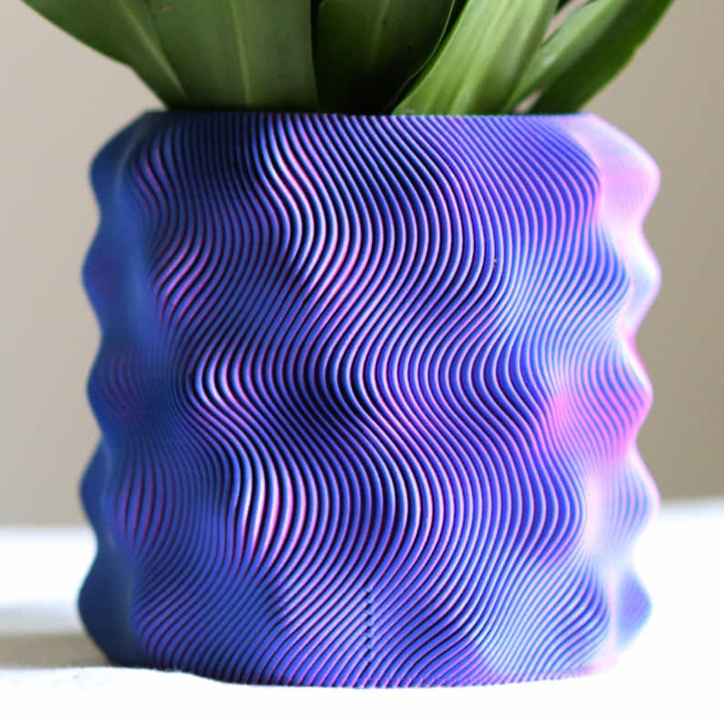 Puede incluir: Una maceta azul y morada con un patr&oacute;n ondulado y una planta en su interior. La maceta tiene un dise&ntilde;o texturizado &uacute;nico con l&iacute;neas verticales que crean una apariencia de onda. La planta tiene hojas largas y verdes.