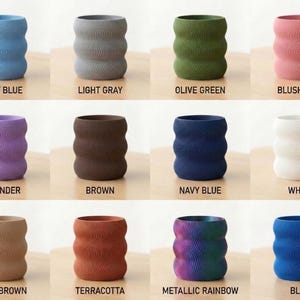 Op de afbeelding: Een verzameling keramische vazen in verschillende kleuren, waaronder lichtblauw, lichtgrijs, olijfgroen, blush roze, lavendel, bruin, marineblauw, wit, lichtbruin, terracotta, metallic regenboog en blauw. Elke vaas heeft een golvend, getextureerd ontwerp.
