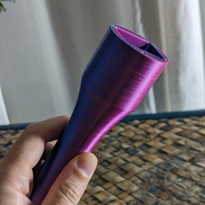 May include: A 3D-printed object with a gradient of blue and purple hues. The object has a cylindrical shape with a flared base and a rectangular opening at the top. The surface has visible layer lines from the printing process.