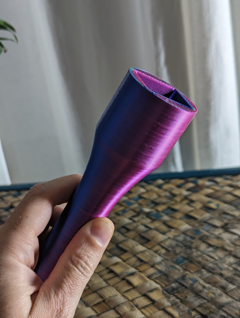 May include: A 3D-printed object with a gradient of blue and purple hues. The object has a cylindrical shape with a flared base and a rectangular opening at the top. The surface has visible layer lines from the printing process.