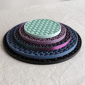 Op de afbeelding: Een stapel van vijf ronde onderzetters in verschillende kleuren: zwart, blauw, paars, blauwgroen en groen. Elke onderzetter heeft een golvend, open ontwerp. De onderzetters zijn van groot naar klein gerangschikt, waardoor een gelaagd effect ontstaat.