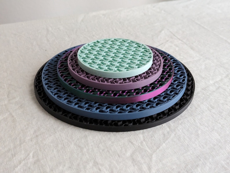 Op de afbeelding: Een stapel van vijf ronde onderzetters in verschillende kleuren: zwart, blauw, paars, blauwgroen en groen. Elke onderzetter heeft een golvend, open ontwerp. De onderzetters zijn van groot naar klein gerangschikt, waardoor een gelaagd effect ontstaat.