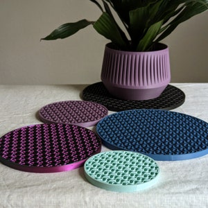 May include: Five round, textured, silicone coasters in different colors: black, purple, blue, pink, and light blue. The coasters are arranged in a circle on a white surface.