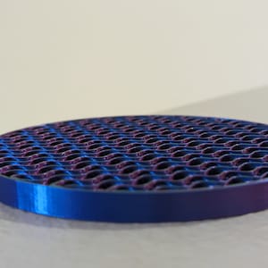 Op de afbeelding: Een ronde, metalen onderzetter met een blauw-paarse gradi&euml;nt. Het oppervlak heeft een herhalend, golfachtig patroon. De onderzetter is ontworpen om oppervlakken te beschermen tegen hitte en vocht.