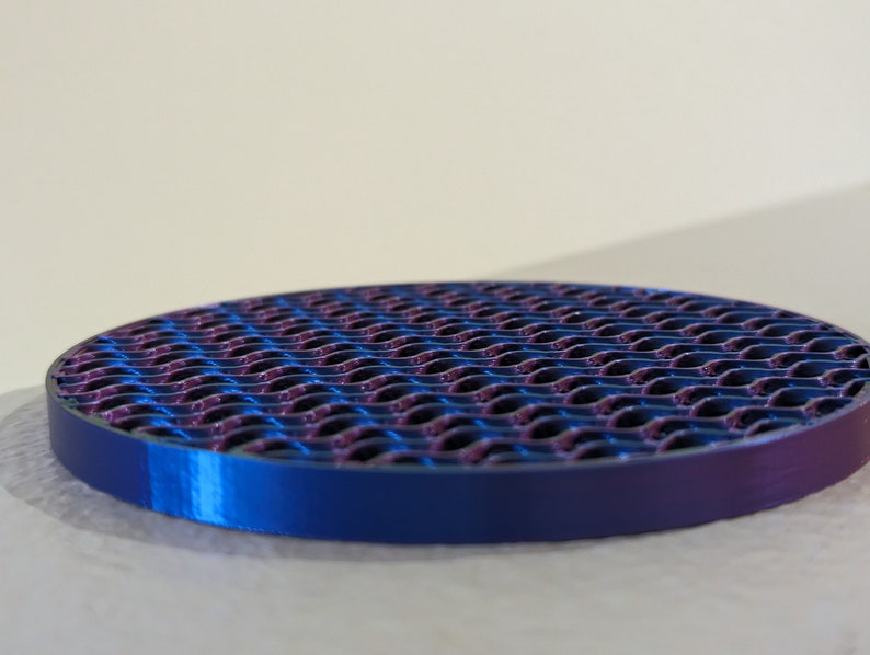 Op de afbeelding: Een ronde, metalen onderzetter met een blauw-paarse gradi&euml;nt. Het oppervlak heeft een herhalend, golfachtig patroon. De onderzetter is ontworpen om oppervlakken te beschermen tegen hitte en vocht.