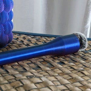 May include: A blue makeup brush with a fluffy, gray brush head. The handle has a metallic sheen. A blue and purple decorative object is in the background. The items are on a woven surface.