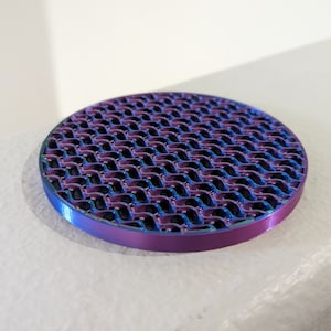 Op de afbeelding: Een ronde, iriserende onderzetter met een golvend, honingraatachtig patroon. De onderzetter heeft een paarse en blauwe kleurovergang, met een gladde, afgeronde rand. Geschikt voor interieurdecoratie.