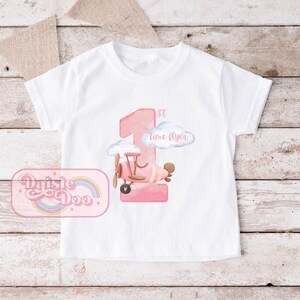 May include: White t-shirt with a pink number one, a pink airplane, and the text "1st time flyer".