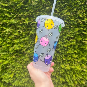 May include: A clear plastic tumbler with a grey background and a repeating pattern of colourful cartoon apples with faces. The apples are yellow, pink, blue, and green. The tumbler has a black lid and a clear straw.
