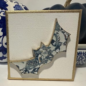 May include: A white canvas with a gold border features a blue and white bat-shaped design. The bat is made of paper with a blue and white floral pattern.