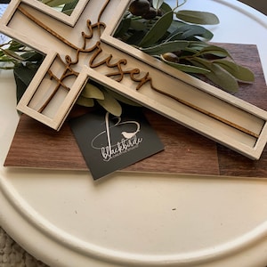 May include: White wooden cross with the words "He is risen" written in cursive on it. The cross is sitting on a wooden surface with a small business card that says "blackbirde creative studio".