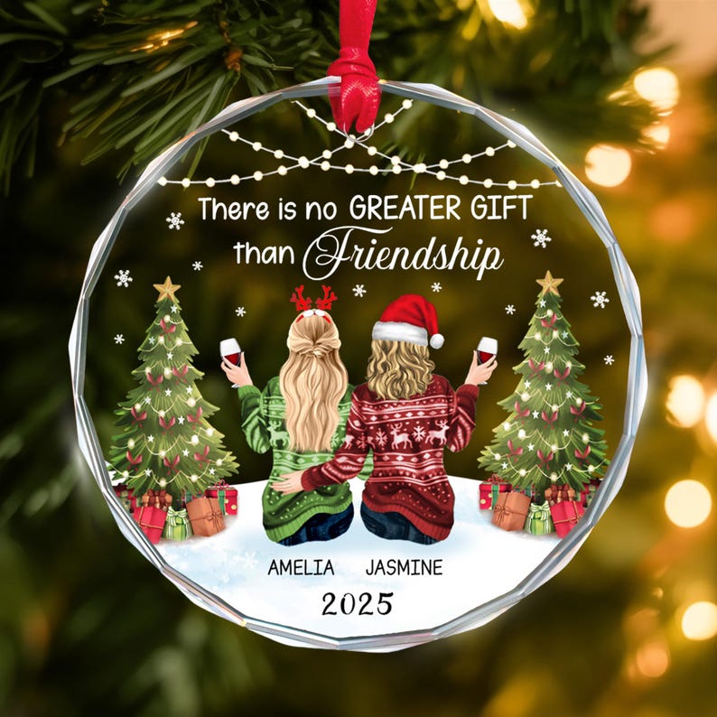 May include: A clear, round Christmas ornament with a red ribbon. The ornament features two figures in sweaters, holding wine glasses, with Christmas trees and the text "There is no GREATER GIFT than Friendship". The names "Amelia" and "Jasmine" are below the figures, with the year "2025".
