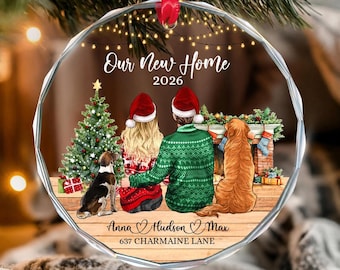 May include: A round, faceted glass Christmas ornament featuring a printed illustration of two people and two dogs sitting by a fireplace. The text reads Our New Home 2026, Anna, Hudson, Max, and 637 CHARMAINE LANE. It hangs from a red ribbon.