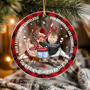 May include: A round ceramic ornament with a red and black buffalo plaid border. The center features an illustration of three people with long hair, wearing sweaters and hats, holding wine glasses. Text reads: Our laughs are limitless, our memories are countless, our friendship is endless. Names Emily, Amelia, and Kayla are printed below.