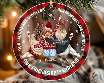 May include: A round ceramic ornament with a red and black buffalo plaid border. The center features an illustration of three people with long hair, wearing sweaters and hats, holding wine glasses. Text reads: Our laughs are limitless, our memories are countless, our friendship is endless. Names Emily, Amelia, and Kayla are printed below.