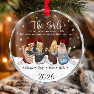 May include: A round, clear glass Christmas ornament featuring an illustration of four people from behind, wearing Santa hats and winter sweaters. The text reads: The Girls, It's not what we have in life, But who we have in our life that matters, Clarissa, Daisy, Grace, Bella, and 2026.