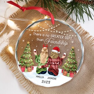 May include: A clear, round Christmas ornament with the text "There is no GREATER GIFT than Friendship." The ornament features two women, Christmas trees, and gifts. The names "AMELIA" and "JASMINE" are below the women, with the year "2025" below the names.