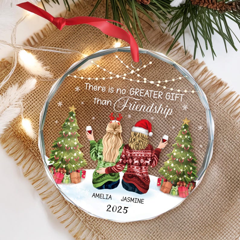 May include: A clear, round Christmas ornament with the text "There is no GREATER GIFT than Friendship." The ornament features two women, Christmas trees, and gifts. The names "AMELIA" and "JASMINE" are below the women, with the year "2025" below the names.