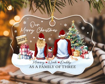 May include: A clear, scalloped-edge Christmas ornament featuring a printed illustration of a family of three sitting in snow. The text reads Our Merry Christmas, 2026, Mommy, Noah, Daddy, and AS A FAMILY OF THREE. Includes a gold hanging cord.