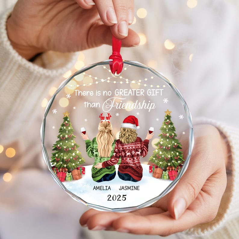 May include: A clear, round Christmas ornament with a red ribbon. The ornament features two figures in sweaters, holding wine glasses, with Christmas trees and the text "There is no GREATER GIFT than Friendship". The names "AMELIA" and "JASMINE" are below the figures, with the year "2025".