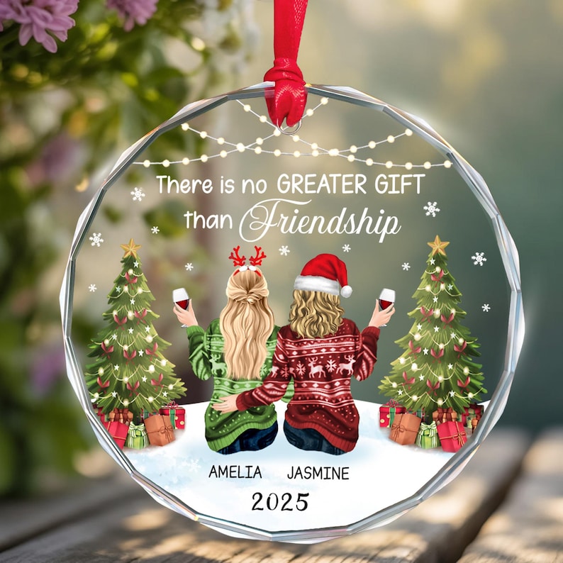 May include: A clear, round Christmas ornament with a red ribbon. The ornament features two figures toasting wine, with Christmas trees and gifts. Text reads "There is no GREATER GIFT than Friendship", with names "AMELIA JASMINE" and the year "2025".