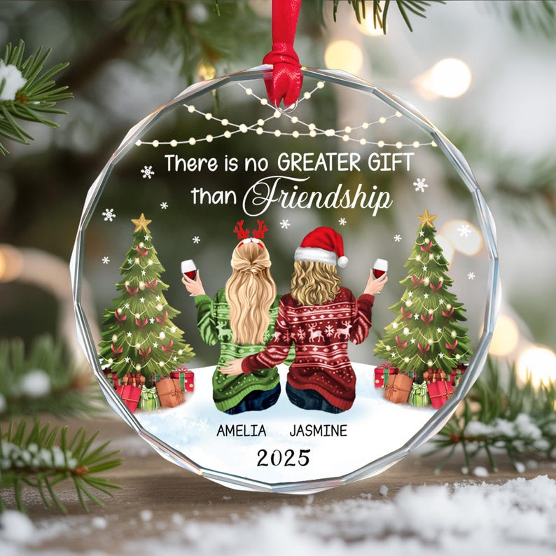 May include: A clear, round Christmas ornament with a red ribbon. The ornament features two women, one in a Santa hat, and the text "There is no GREATER GIFT than Friendship." The names "AMELIA" and "JASMINE" are below the women, with the year "2025".