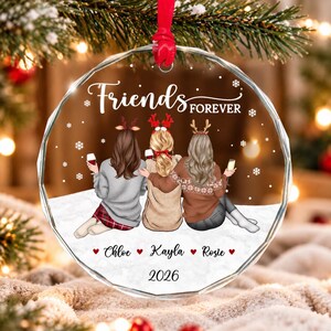 May include: A round, clear glass ornament featuring an illustration of three friends with reindeer antlers, sitting in snow. The text reads Friends FOREVER, Chloe, Kayla, Rosie, and 2026. The friends wear sweaters and hold wine glasses.
