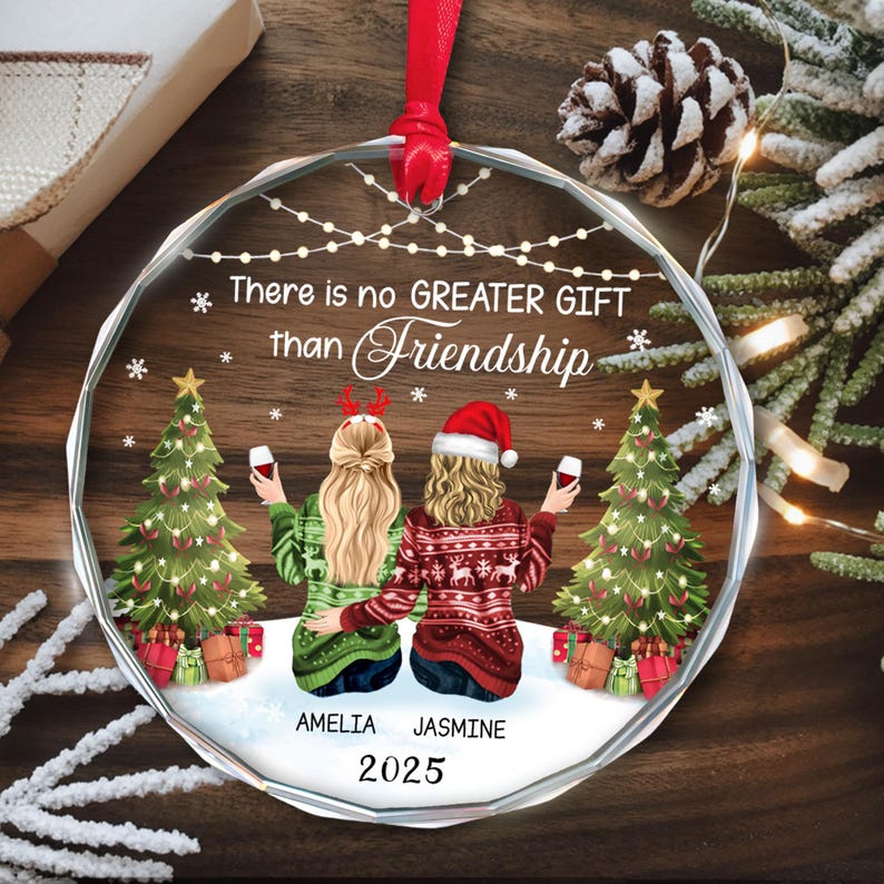May include: A clear, round Christmas ornament with a red ribbon. The ornament features two women, Christmas trees, and the text "There is no GREATER GIFT than Friendship." The names "AMELIA" and "JASMINE" are below the women, with the year "2025" below the names.