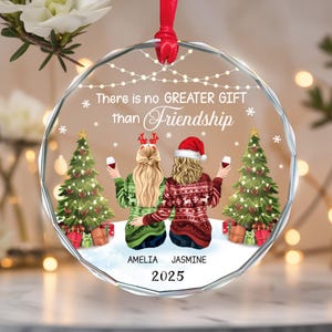 May include: A clear, round Christmas ornament with a red ribbon. The ornament features two figures in sweaters, holding wine glasses, with Christmas trees and the text "There is no GREATER GIFT than Friendship". The names "AMELIA" and "JASMINE" are below the figures, with the year "2025".