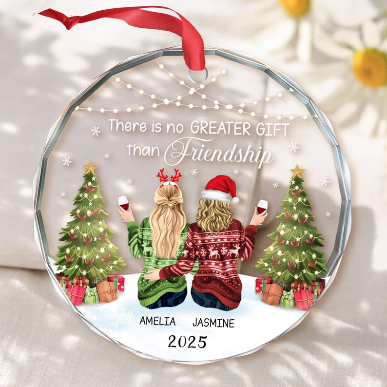 May include: A clear, round Christmas ornament with a red ribbon. The ornament features two figures in festive sweaters, holding wine glasses, with Christmas trees and the text "There is no GREATER GIFT than Friendship". The names "AMELIA" and "JASMINE" are below the figures, with the year "2025".