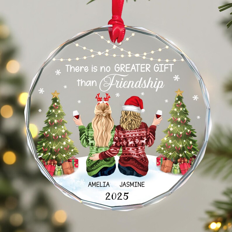 May include: A clear, round Christmas ornament with a red ribbon. The ornament features two figures in sweaters, holding wine glasses, with Christmas trees and the text "There is no GREATER GIFT than Friendship". The names "AMELIA" and "JASMINE" are below the figures, with the year "2025".