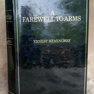 May include: A hardcover book titled "A Farewell to Arms" by Ernest Hemingway. The book has a dark green cover with gold lettering and trim. The title and author's name are prominently displayed on the front. Part of "The John Player Special Collection" is visible.