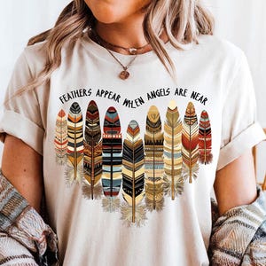 May include: A white t-shirt with a graphic of colorful feathers and the text "Feathers Appear When Angels Are Near".