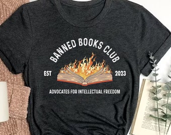 Puede incluir: Una camiseta gris oscuro con el texto "Banned Books Club Est 2023 Advocates for Intellectual Freedom" y un gráfico de un libro en llamas.