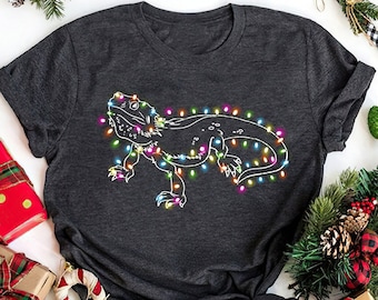 May include: A dark gray t-shirt with a white outline of a bearded dragon decorated with colorful Christmas lights. The shirt is surrounded by Christmas decorations, including presents, candy canes, and ornaments.