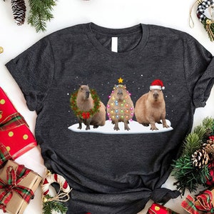 May include: A dark gray t-shirt with a Christmas-themed design featuring three capybaras. One capybara is wearing a wreath, another is decorated as a Christmas tree with lights, and the third is wearing a Santa hat.