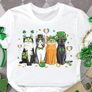 May include: White t-shirt with a graphic of four cats dressed in St. Patrick's Day attire. The cats are wearing hats, shamrock glasses, and bow ties. The graphic also includes shamrocks, gold coins, and balloons.