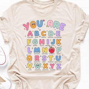 May include: A beige t-shirt featuring a colourful alphabet design. Each letter is surrounded by a star and has a positive word associated with it. The words are: Accepting, Brave, Creative, Dedicated, Encouraging, Friendly, Genuine, Helpful, Inspiring, Joyful, Kind, Loving, Motivated, Nurturing, Optimistic, Patient, Quirky, Respectful, Smart, Thoughtful, Understanding, Valued, Wonderful, Xenodochial, Youthful, Zealous. The shirt has the text "You Are" at the top.