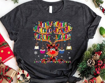 May include: Dark gray t-shirt featuring a reindeer design with antlers made of colorful books. The reindeer has a red nose and is wearing a plaid pattern. The design includes hanging books and Christmas lights, perfect for book lovers during the holiday season.