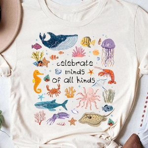 Puede incluir: Camiseta color crema con un diseño colorido de criaturas marinas, incluyendo una ballena, peces y un pulpo. El texto "celebrate minds of all kinds" está en el centro, rodeado de ilustraciones de vida marina.