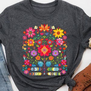 May include: A dark grey t-shirt with a colourful floral design. The design features a variety of flowers in shades of red, pink, yellow, and blue. The shirt is paired with blue jeans and brown shoes.
