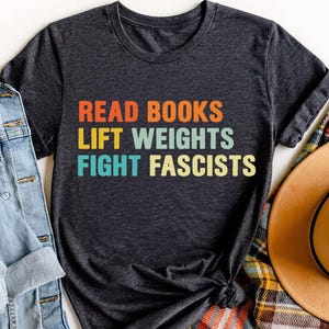 May include: Dark gray t-shirt with the text "READ BOOKS LIFT WEIGHTS FIGHT FASCISTS" in orange, blue, yellow, and green. The shirt is knotted at the bottom and is next to a denim jacket and a hat.