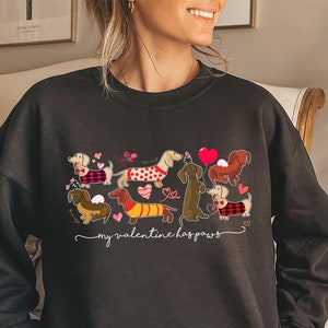 Puede incluir: Una sudadera negra de manga larga con un estampado gr&aacute;fico de seis perros salchicha con diversos atuendos, rodeados de corazones rosas y rojos y el texto "my valentine has paws" escrito en letra cursiva blanca sobre el pecho.