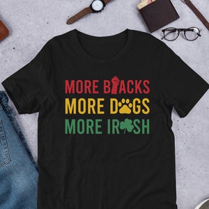 以下が含まれることがあります： 黒のTシャツに「More Blacks, More Dogs, More Irish」という文字が赤、黄、緑で書かれています。