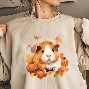 Peut inclure: Un sweat-shirt beige avec un imprimé graphique d'un cochon d'Inde brun et blanc entouré de citrouilles orange et de feuilles d'automne.