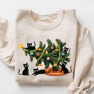 Pode incluir: Camisola bege com um design natalino. Apresenta uma árvore de Natal decorada com enfeites e luzes, rodeada por vários gatos pretos. Os gatos interagem com a árvore, criando uma cena festiva e divertida.
